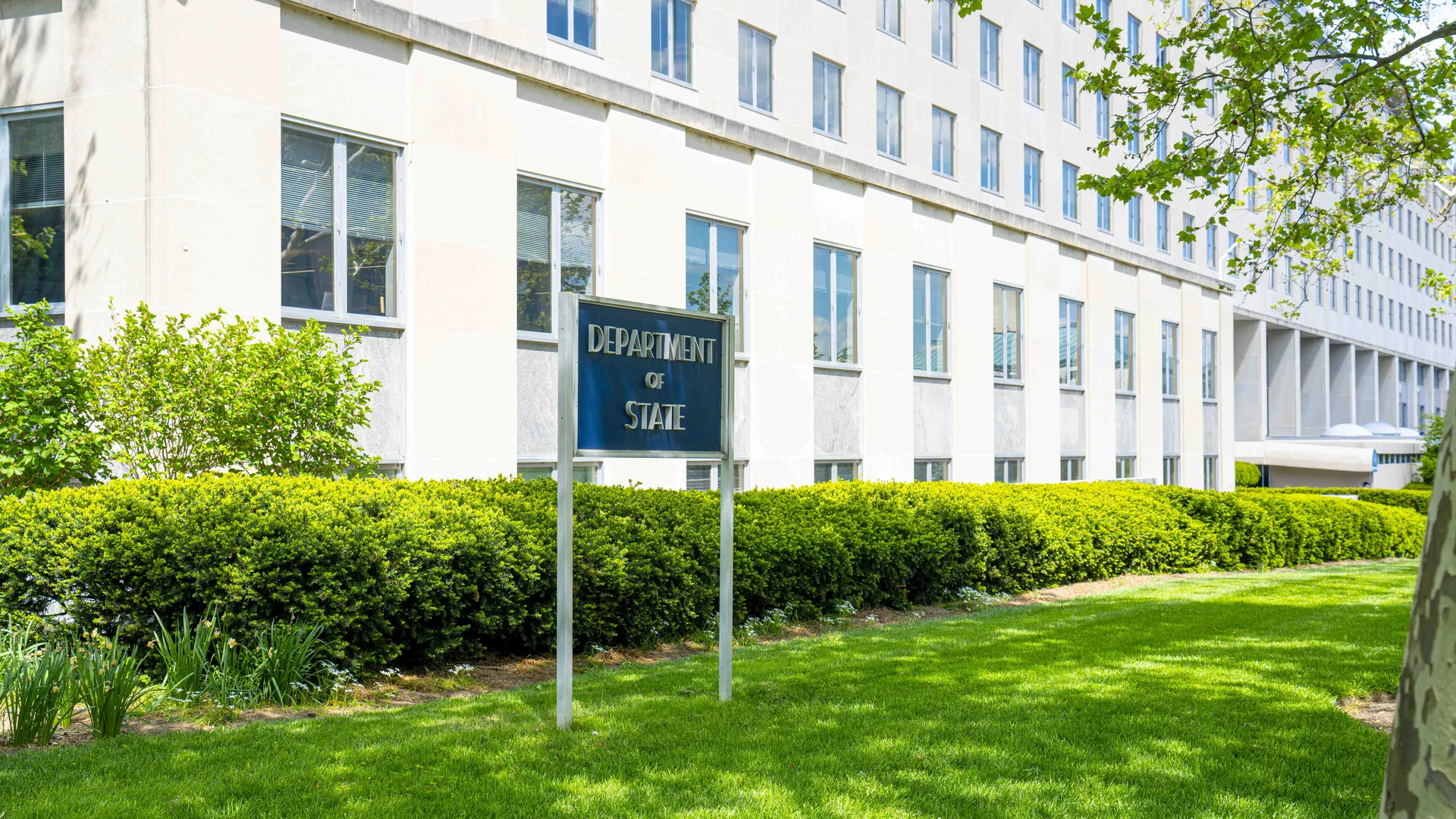 A blue "Department of State" sign on the lawn in front of the Harry S. Truman Building, the State Department headquarters in Washington, D.C., in spring.