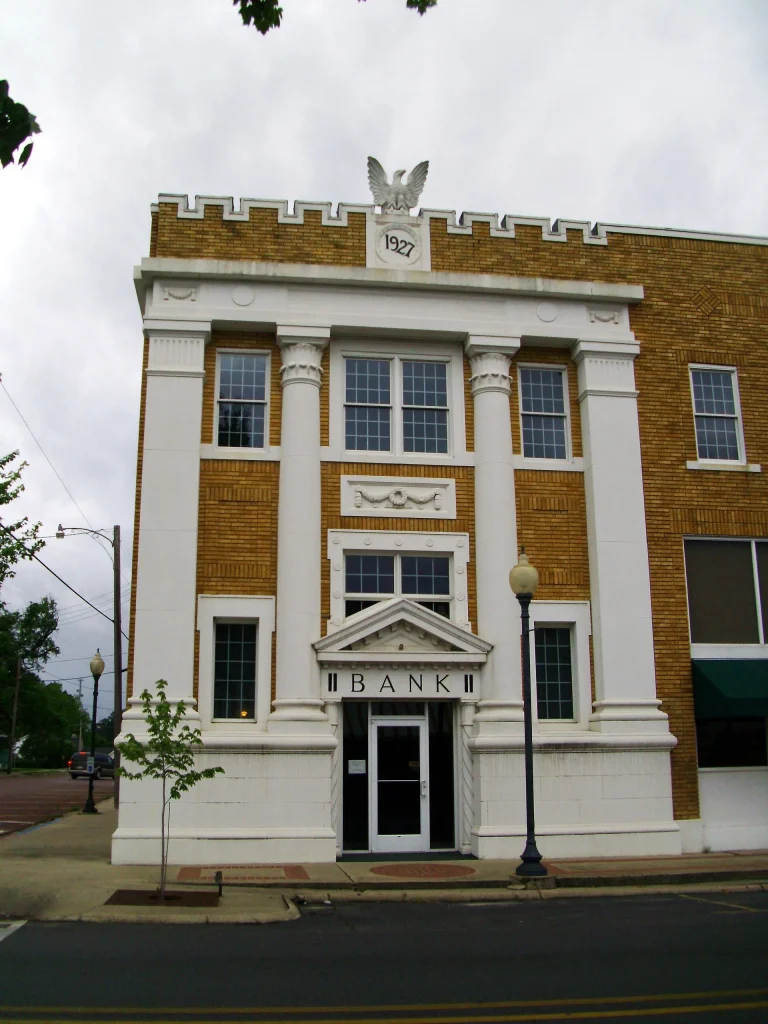 Narrow two-story bank building with a white stone front, columns, a carved eagle on top and the word BANK over the door, beside a tan brick building.