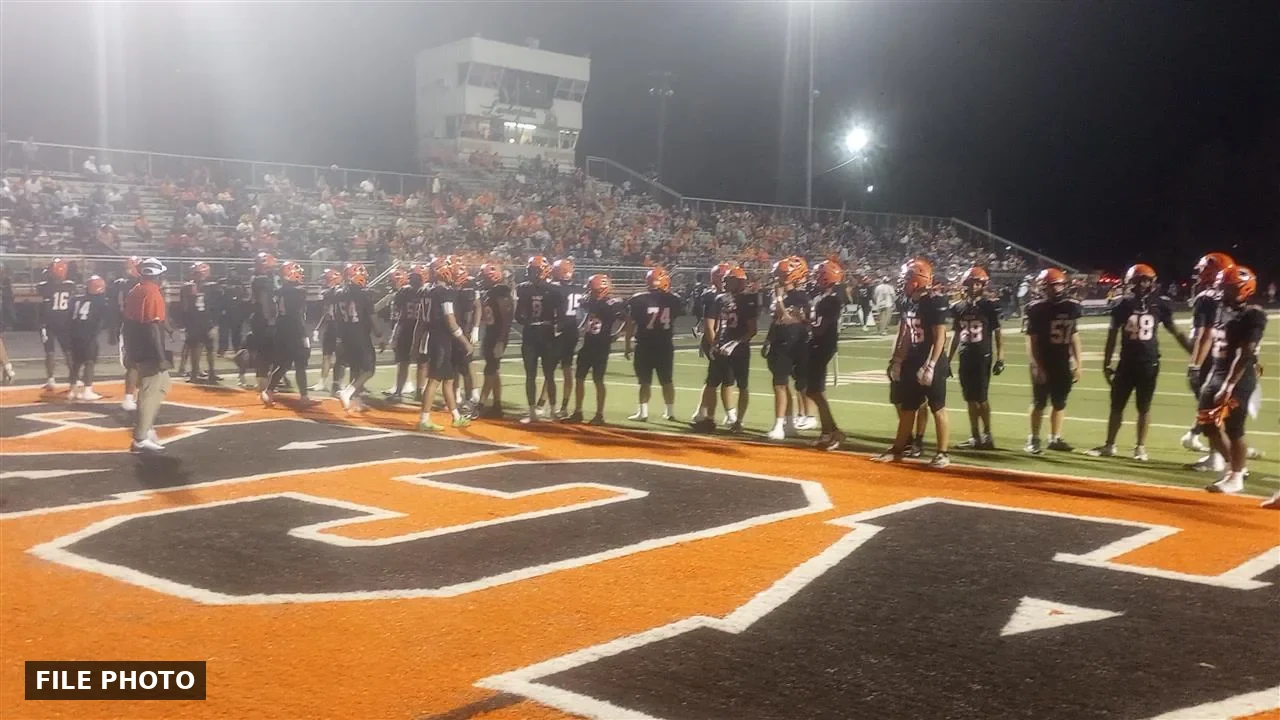 Football players in black jerseys and orange helmets standing along a sideline beside a large orange end-zone letter at night, with a crowded grandstand and press box behind them, marked File Photo