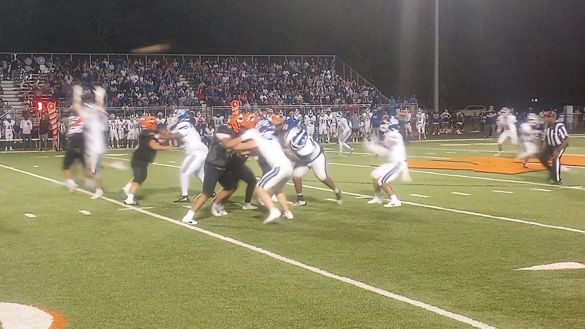 Warren players in black jerseys and orange helmets and Monticello players in white jerseys collide at the line of scrimmage under stadium lights, with an official nearby.