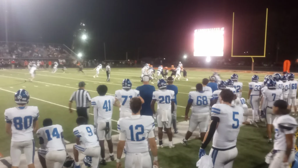 Monticello players in white jerseys watch a play from the sideline at night, with the Lumberjacks scoreboard and yellow goalposts in the background.