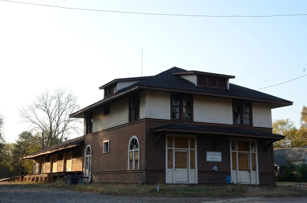 Two-story brick and stucco former railway station with a wide hipped roof and a long covered platform along one side, next to an empty lot.