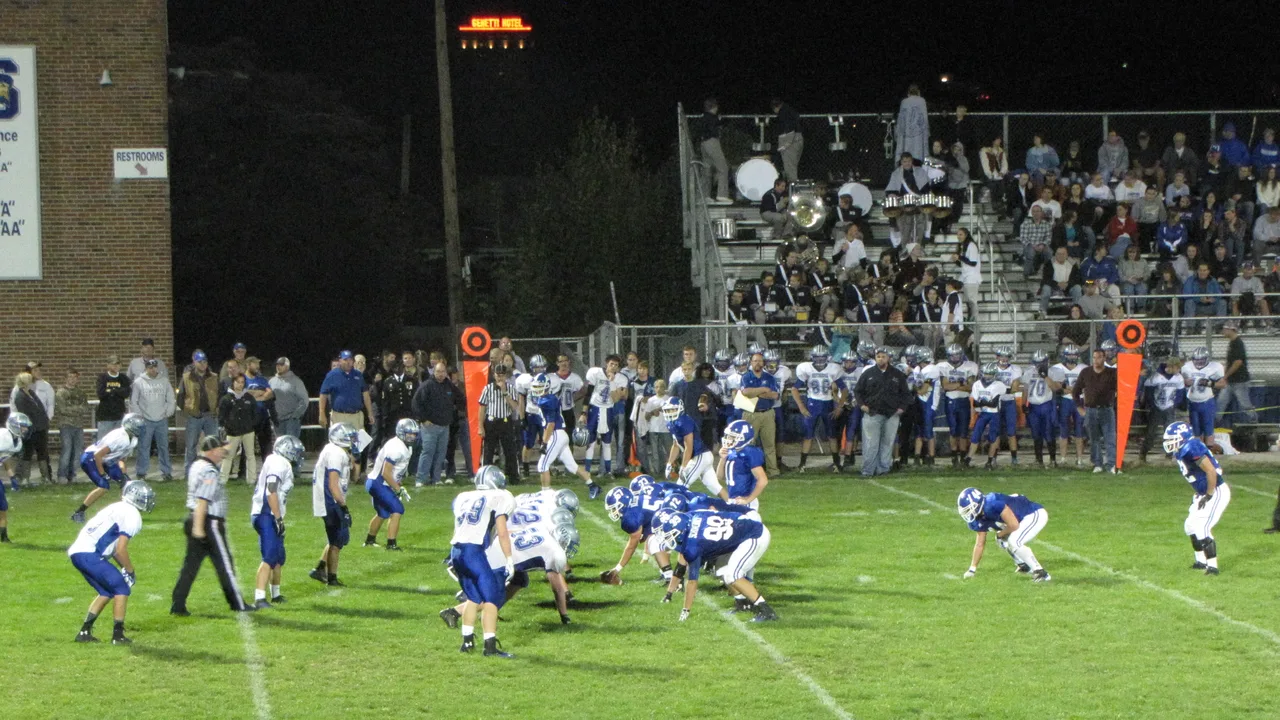 High school football teams in blue and white jerseys line up at the line of scrimmage during a night game in front of packed bleachers
