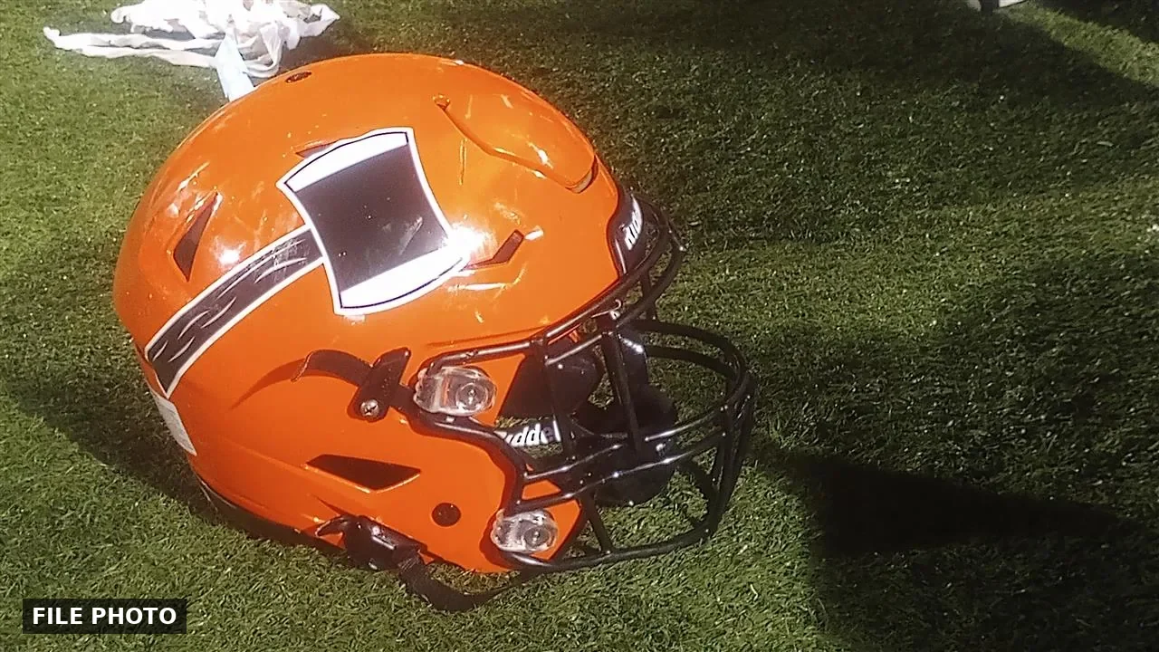 Orange football helmet with a black facemask and a black-and-white side decal lying on green artificial turf, marked File Photo