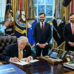 President Donald Trump signs an executive order at the Resolute Desk in the Oval Office as Vice President JD Vance and another official stand behind him, March 16, 2026.