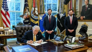 President Donald Trump signs an executive order at the Resolute Desk in the Oval Office as Vice President JD Vance and another official stand behind him, March 16, 2026.