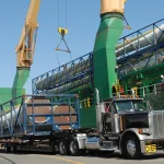 Long white wind turbine blades in blue steel frames being unloaded from a cargo ship onto an oversize-load truck at a port, with green cranes overhead.