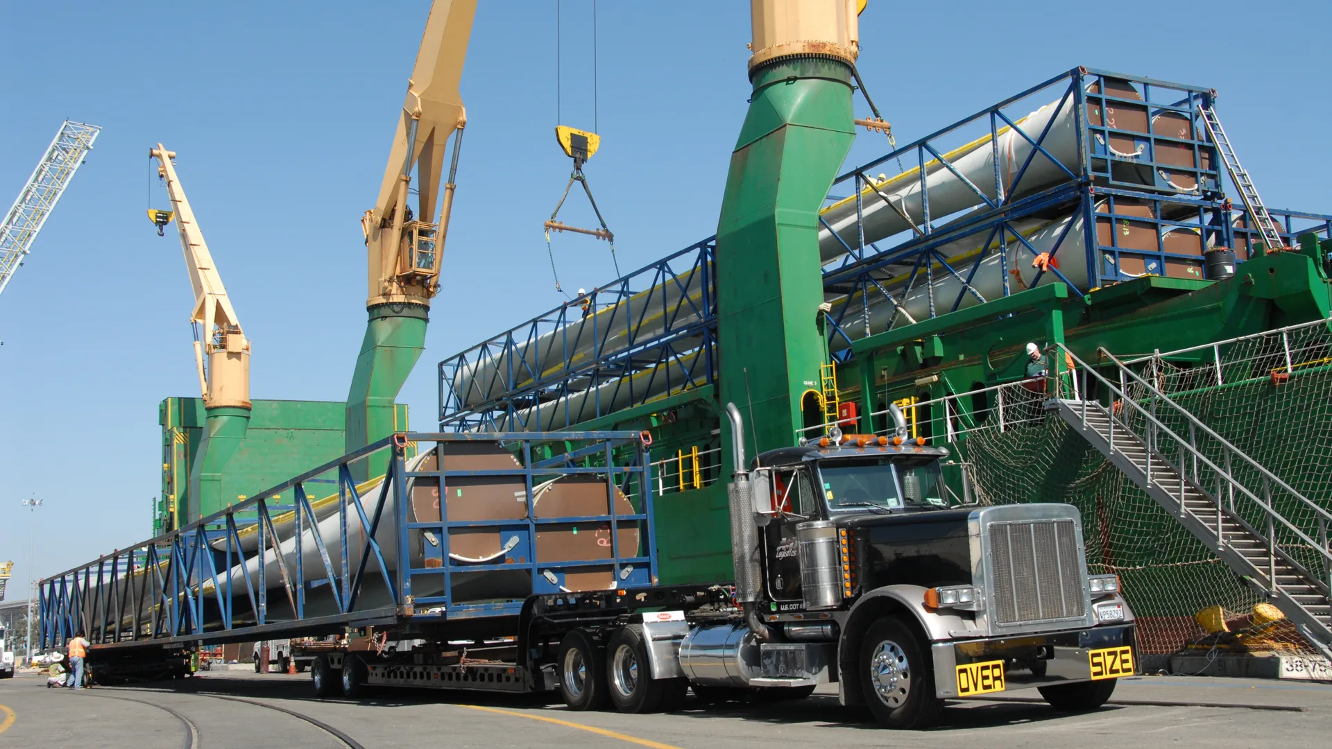 Long white wind turbine blades in blue steel frames being unloaded from a cargo ship onto an oversize-load truck at a port, with green cranes overhead.