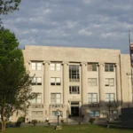 The Drew County Courthouse in Monticello, a three-story stone building with tall columns, behind trees under a cloudy sky.
