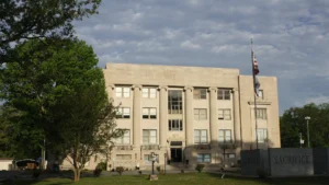 The Drew County Courthouse in Monticello, a three-story stone building with tall columns, behind trees under a cloudy sky.