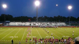 A football game kicks off under stadium lights at dusk, seen from the stands.