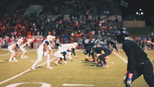 Two football teams set at the line of scrimmage under night lights in front of a full grandstand.
