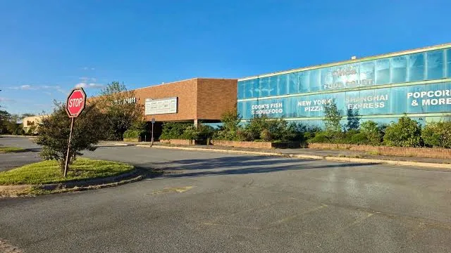 Exterior of the Pine Bluff Mall food court entrance on a clear day: a glass front with signs for Cook's Fish & Soulfood, New York Pizza, Shanghai Express and Popcorn & More, next to a brown brick section, with a stop sign and empty pavement in the foreground.