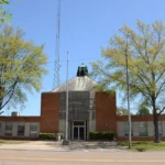 The front of the Crossett Municipal Building in Arkansas, a one-story brick building with a central entrance, surrounded by trees and lawn.