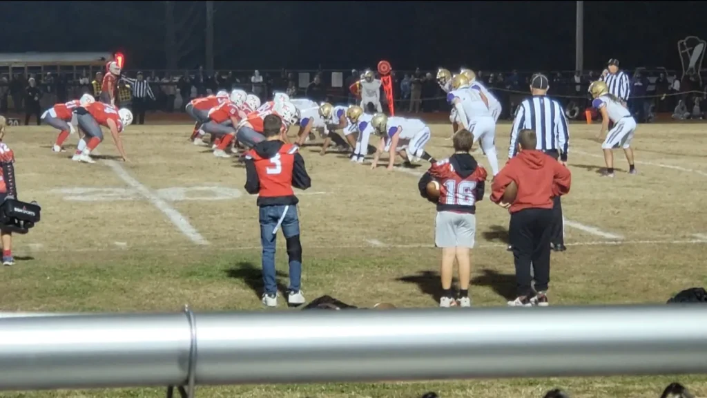Two high school football teams, one in red and one in white and gold, lined up at the line of scrimmage under stadium lights with officials and ball boys on the sideline.