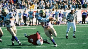 A ball carrier in a white and purple uniform runs past a fallen defender in red while a referee follows, with a packed grandstand behind.