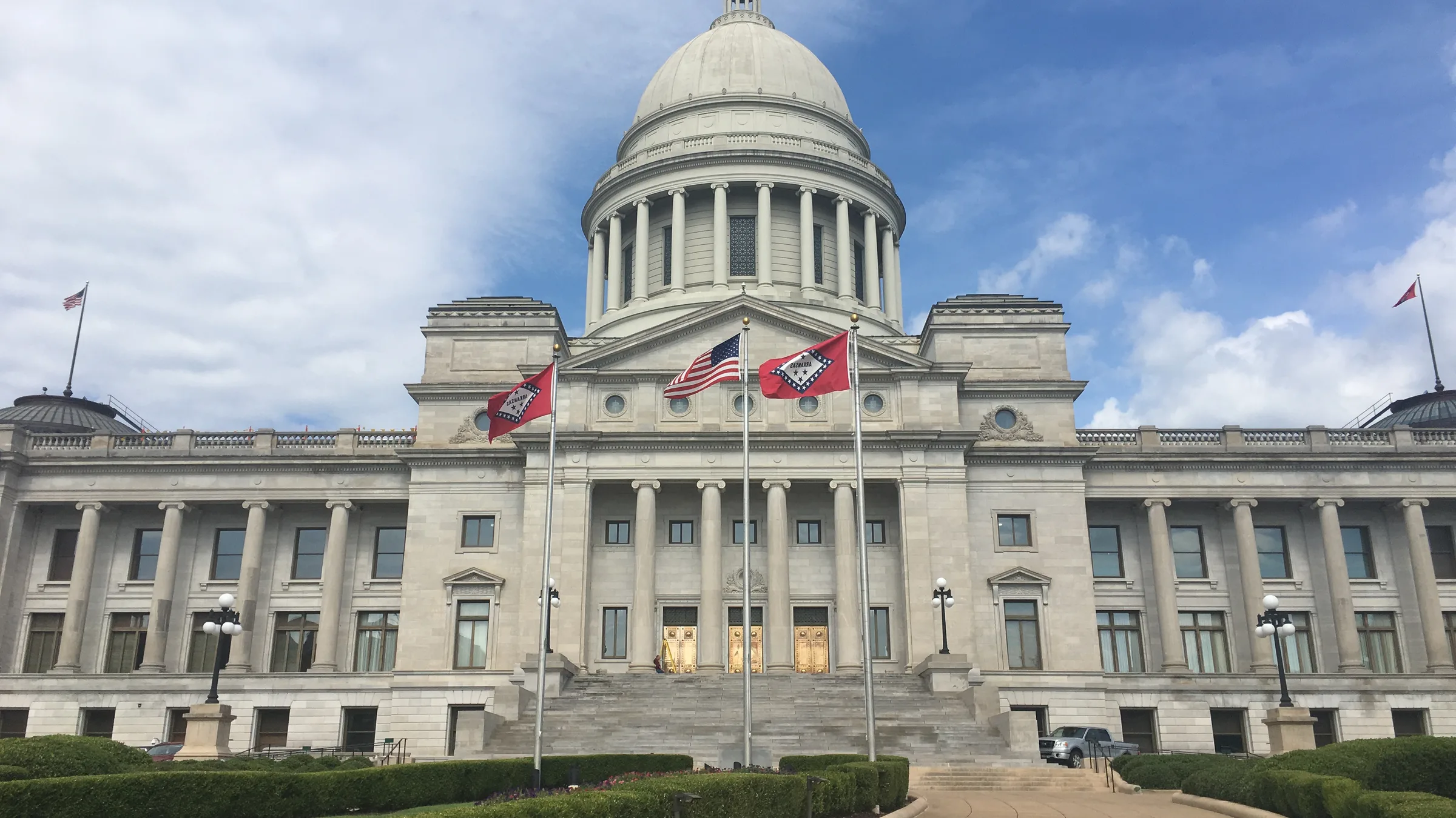 The Arkansas State Capitol in Little Rock, a white neoclassical building with a central dome and U.S. and Arkansas flags in front.