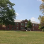 A two-story red-brick building with white window trim framed by large trees on the University of Arkansas at Monticello campus.