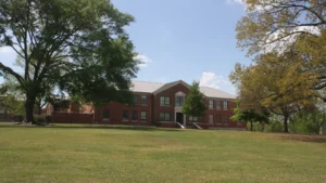 A two-story red-brick building with white window trim framed by large trees on the University of Arkansas at Monticello campus.