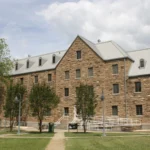 A three-story stone academic building with a gabled metal roof behind a green lawn on the University of Arkansas at Monticello campus.