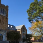 The historic Music Building on the University of Arkansas at Monticello campus, a tan brick building with a tower beside a large tree under a clear blue sky.
