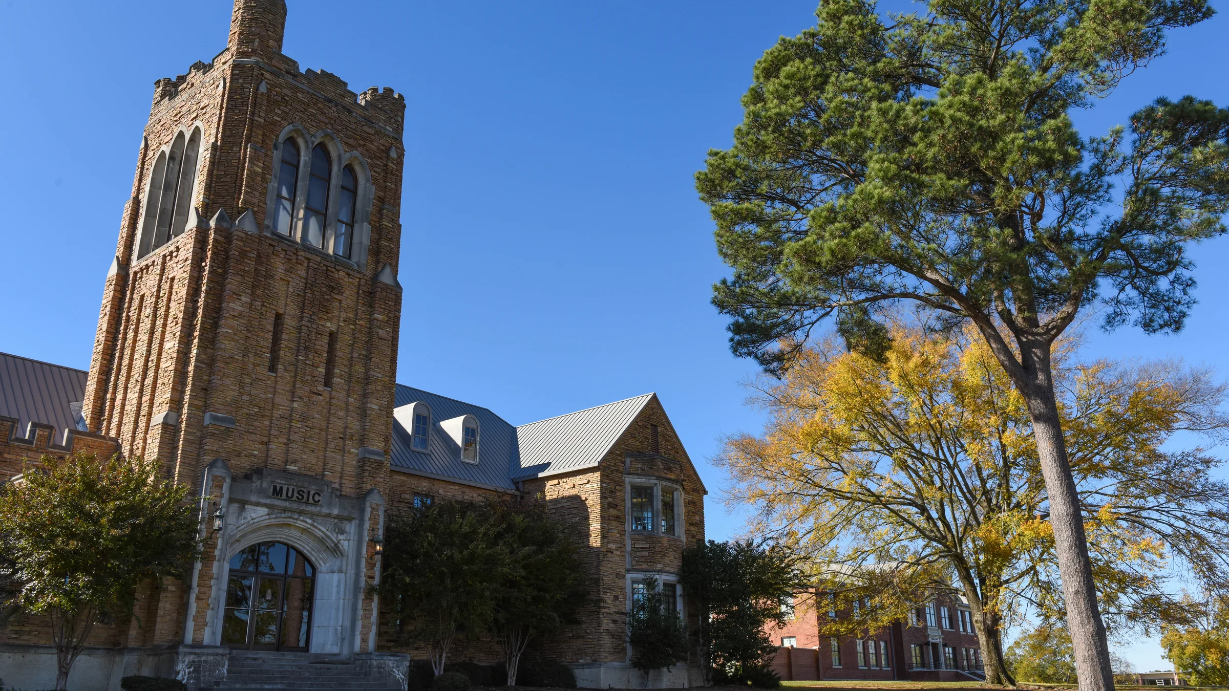 The historic Music Building on the University of Arkansas at Monticello campus, a tan brick building with a tower beside a large tree under a clear blue sky.